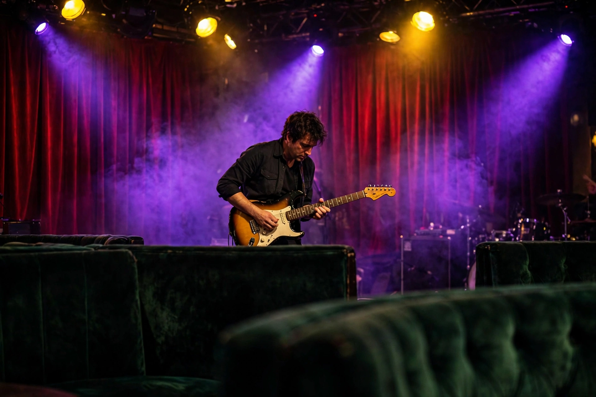 Blues club stage under violet and yellow spotlights: a guitarist playing a sunburst Stratocaster against a red curtain, green velvet booths in the foreground